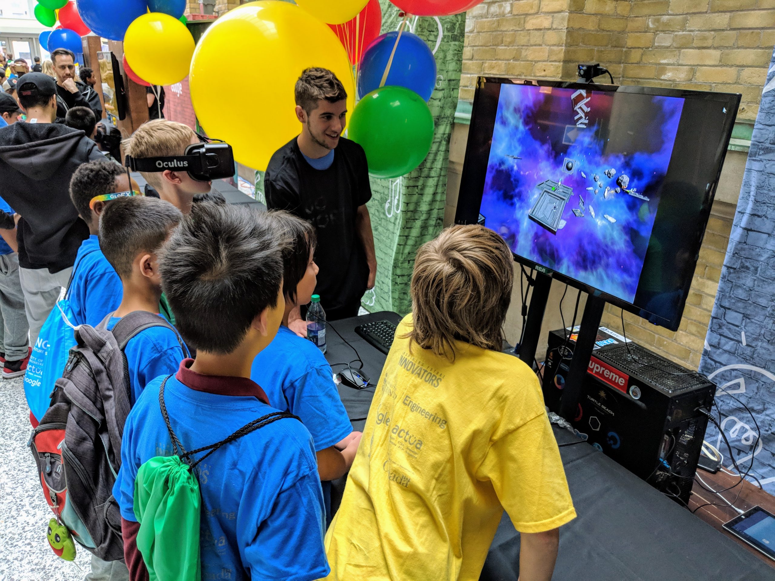 Photo of a public event for at-risk middle school youth, with half a dozen students crowding around while one of their friends plays a virtual reality game on a large-screen display. The game features a main character who has to solve coding problems in order to advance from platform to platform. One of the developers is watching from the side and providing encouragement from the student playing the game.