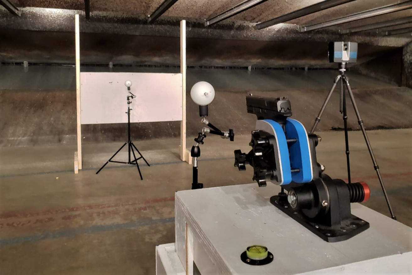 Photo at a shooting range of a semi-automatic handgun, held in a gun rest on a table, and aimed a section of drywall. Two spheres on tripods are placed between the gun and the drywall panel. A 3D laser scanner is placed to the side to capture the scene.