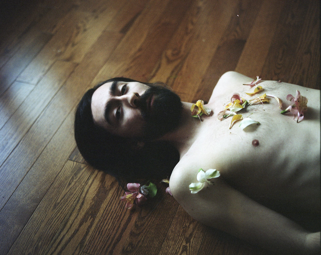 Image of shirtless man with dark hair and beard and light skin with flower petals on top of him. Moody vibes.